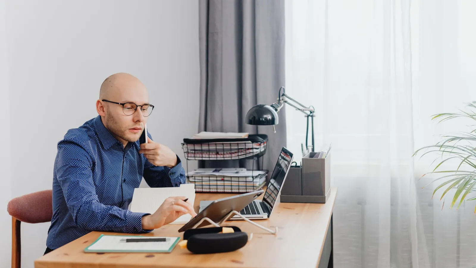 Business owner at a home office desk working on a laptop and tablet with a phone to his ear