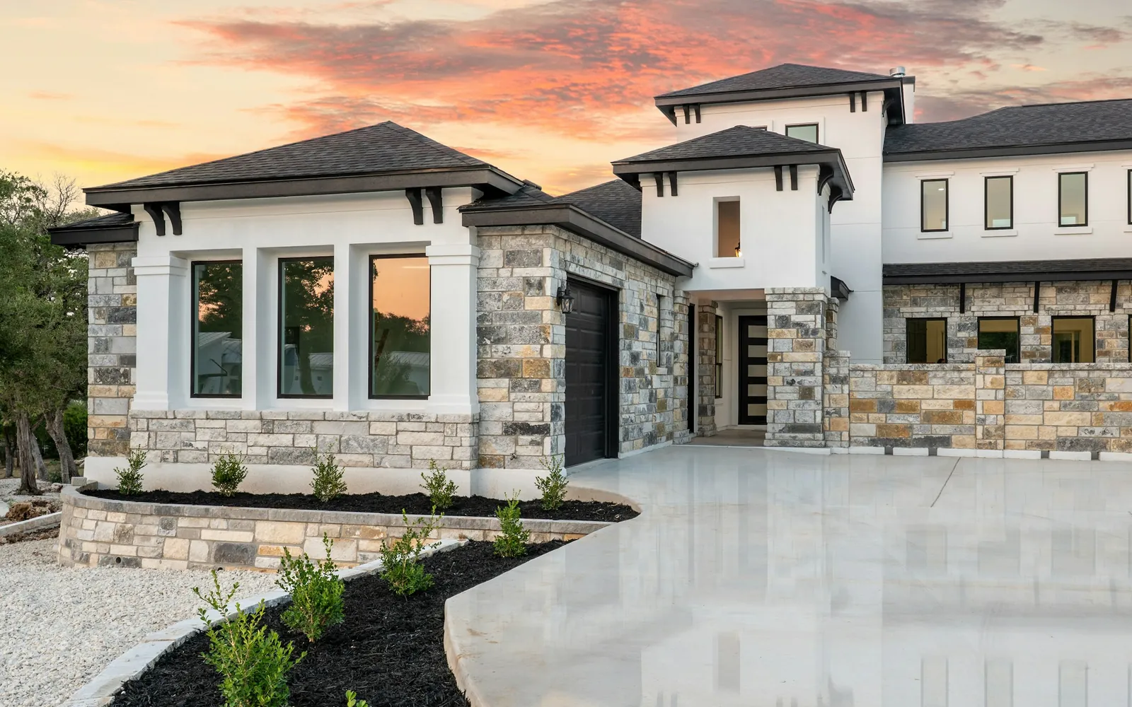 A house with light stone and white stucco walls, dark gray asphalt shingle roofs, a dark garage door, a concrete driveway, and young plants in mulch beds at sunset