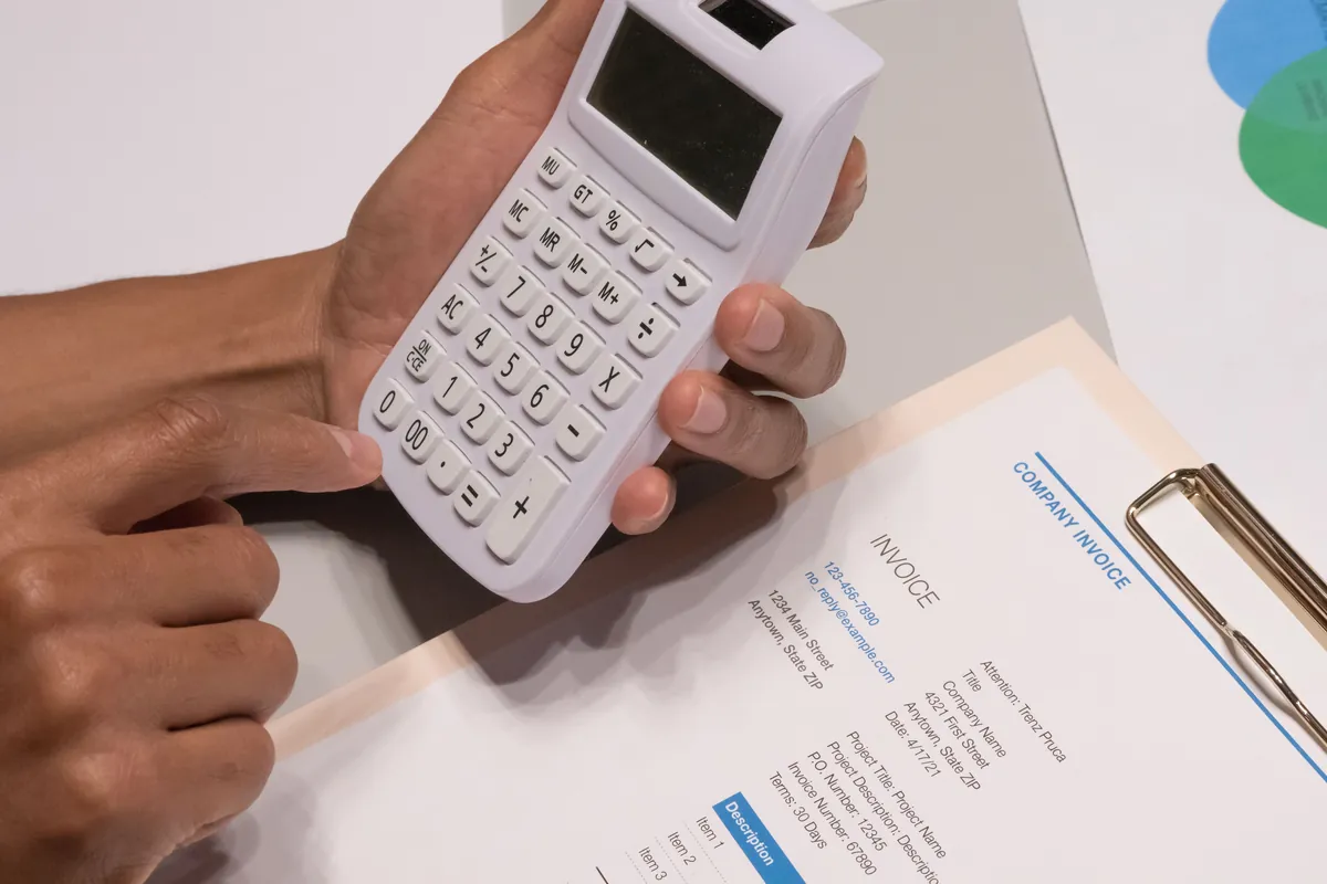 Hands holding a white calculator over a company invoice on a clipboard